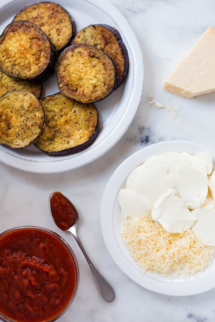 An overhead image of a kitchen counter with all the ingredients for the Baked Eggplant Parmesan including roasted eggplant, homemade tomato sauce, mozzarella and grated parmesan.