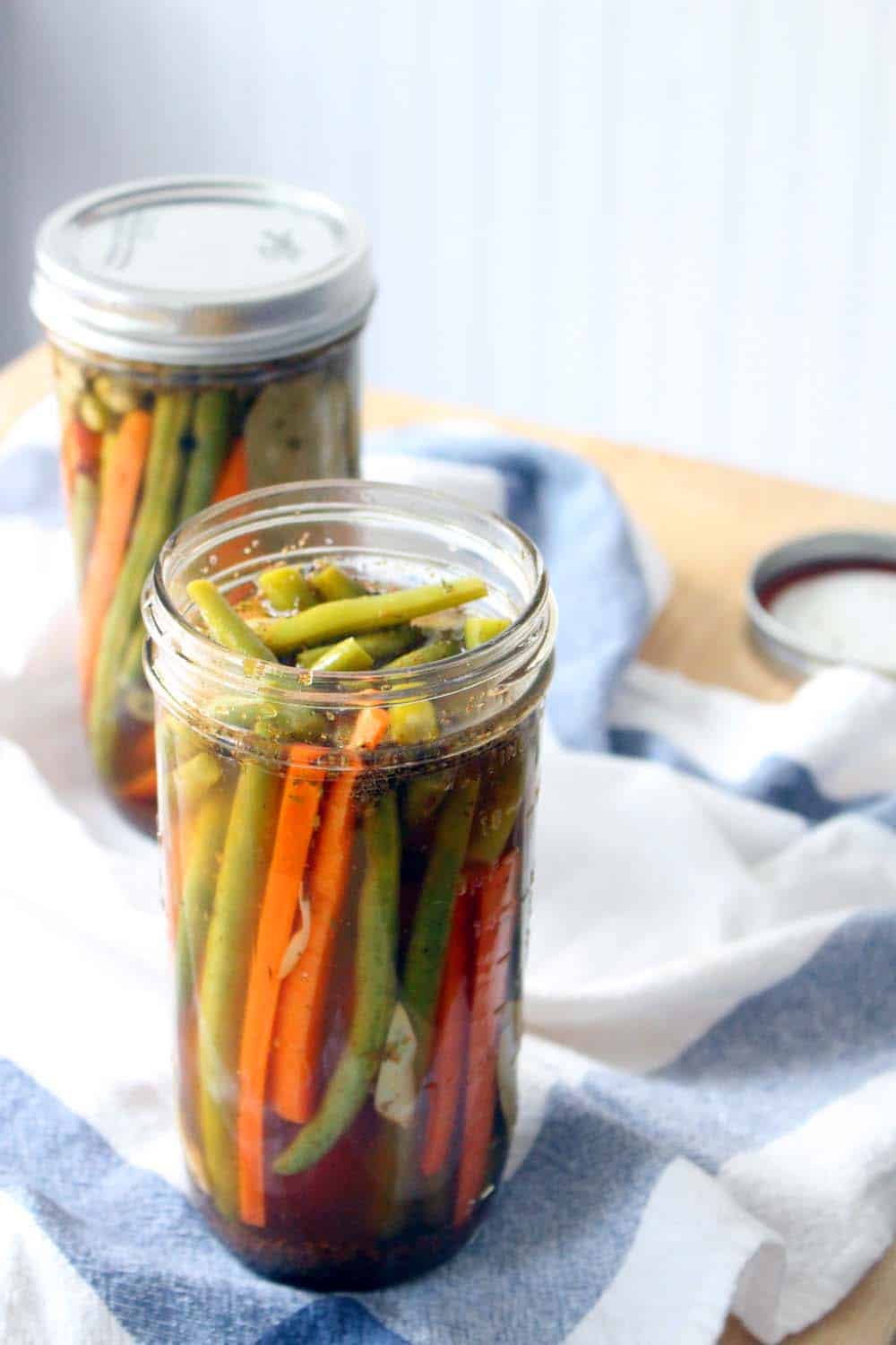 Two tall glass jars holding pickled green beans and carrots. One is in the foreground with an open top, and one is in the background with the lid on.