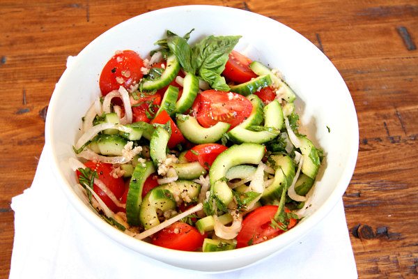 Overhead shot of Tomato Cucumber and Basil Salad in a white bowl with a white napkin on a wood surface